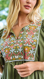 A woman wearing an olive green short-sleeve dress with a colorful floral embroidered yoke, standing on a white porch with a lush green garden background.