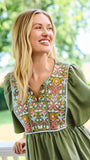 A woman wearing an olive green short-sleeve dress with a colorful floral embroidered yoke, standing on a white porch with a lush green garden background.