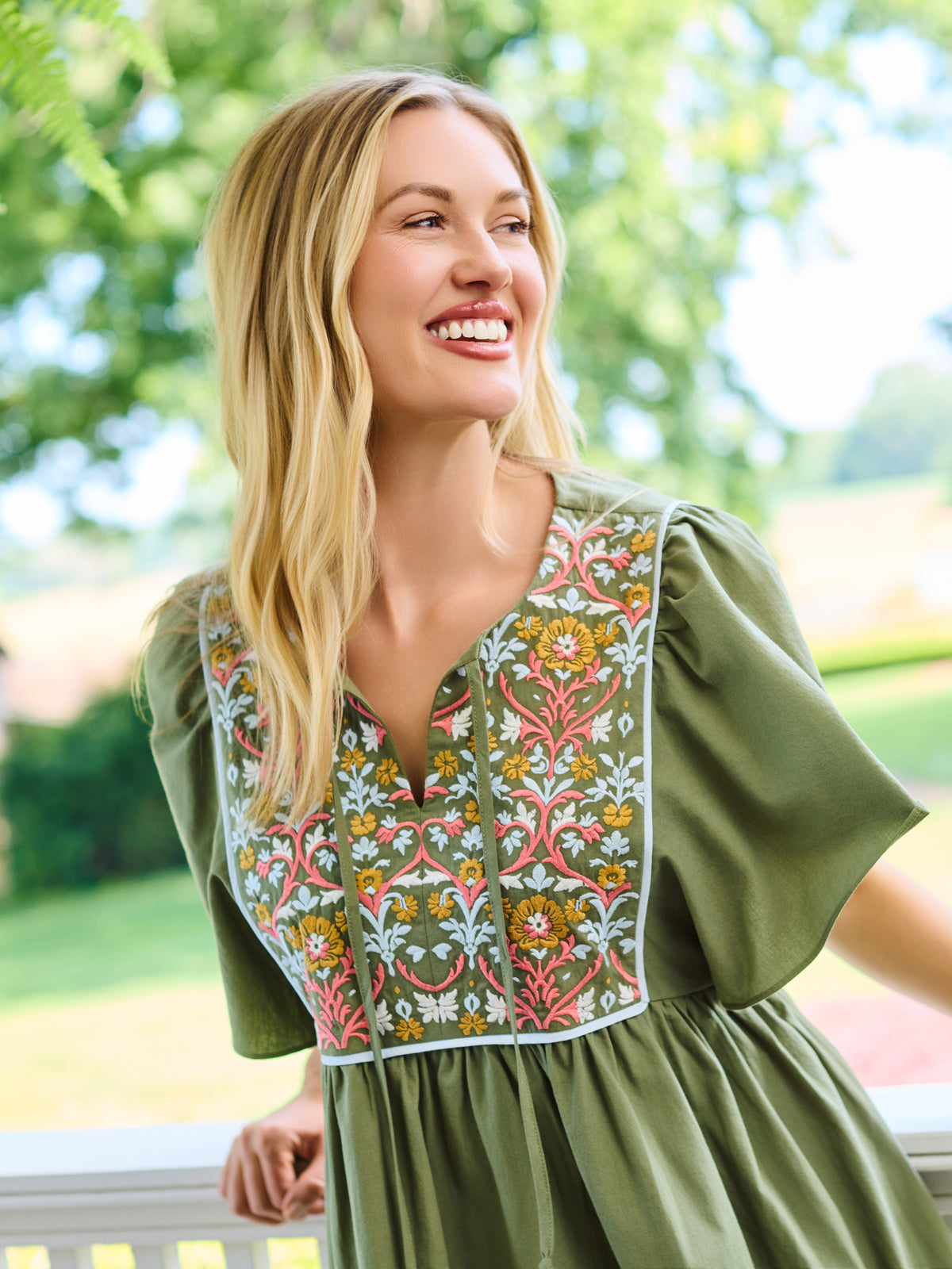 A woman wearing an olive green short-sleeve dress with a colorful floral embroidered yoke, standing on a white porch with a lush green garden background.