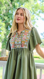 A woman wearing an olive green short-sleeve dress with a colorful floral embroidered yoke, standing on a white porch with a lush green garden background.