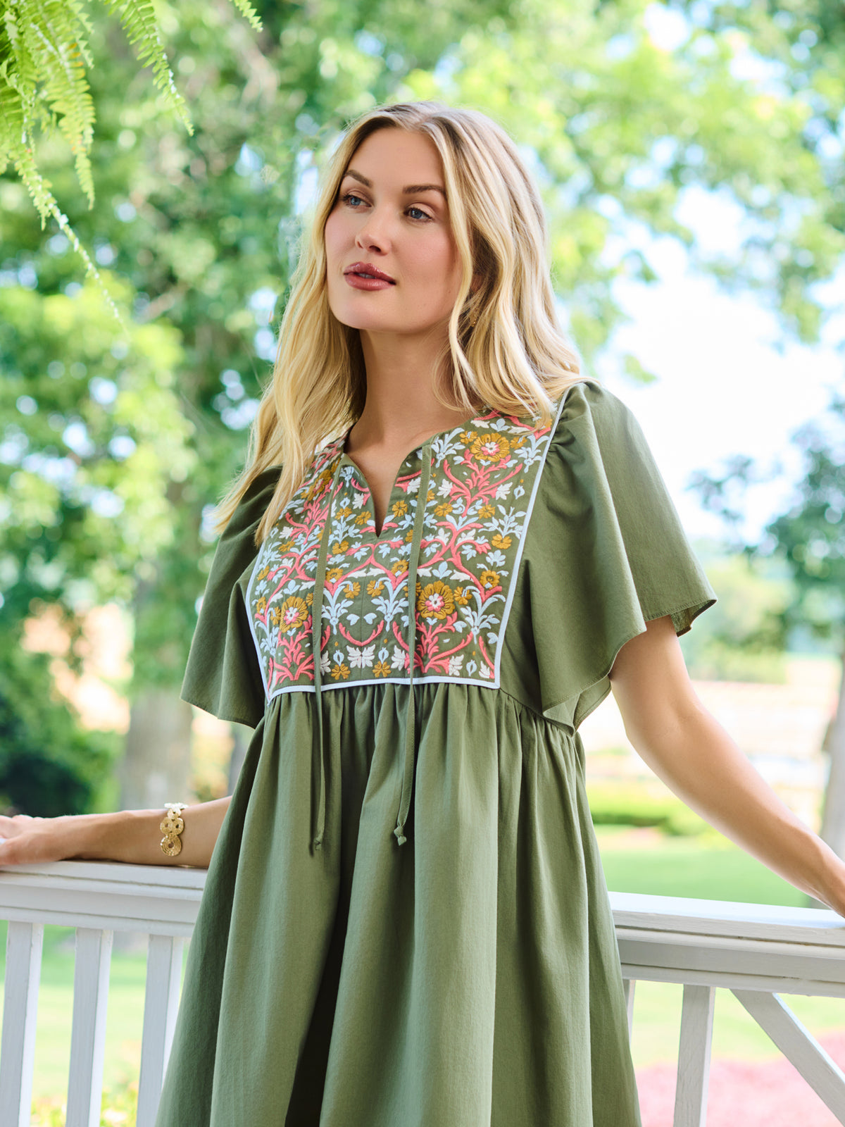 A woman wearing an olive green short-sleeve dress with a colorful floral embroidered yoke, standing on a white porch with a lush green garden background.