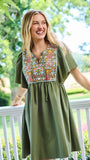A woman wearing an olive green short-sleeve dress with a colorful floral embroidered yoke, standing on a white porch with a lush green garden background.
