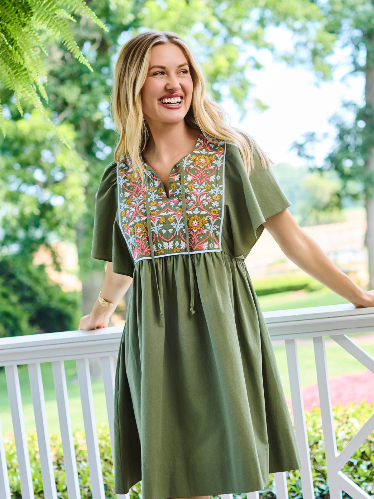 A woman wearing an olive green short-sleeve dress with a colorful floral embroidered yoke, standing on a white porch with a lush green garden background.