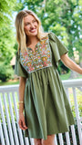 A woman wearing an olive green short-sleeve dress with a colorful floral embroidered yoke, standing on a white porch with a lush green garden background.