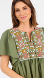 A woman modeling a short-sleeve olive green dress with a colorful floral embroidered yoke and a gathered waist, paired with beige flat shoes against a plain white background.