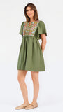 A woman modeling a short-sleeve olive green dress with a colorful floral embroidered yoke and a gathered waist, paired with beige flat shoes against a plain white background.