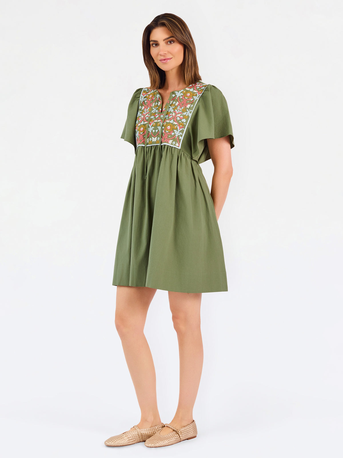 A woman modeling a short-sleeve olive green dress with a colorful floral embroidered yoke and a gathered waist, paired with beige flat shoes against a plain white background.