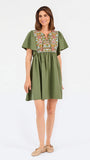 A woman modeling a short-sleeve olive green dress with a colorful floral embroidered yoke and a gathered waist, paired with beige flat shoes against a plain white background.