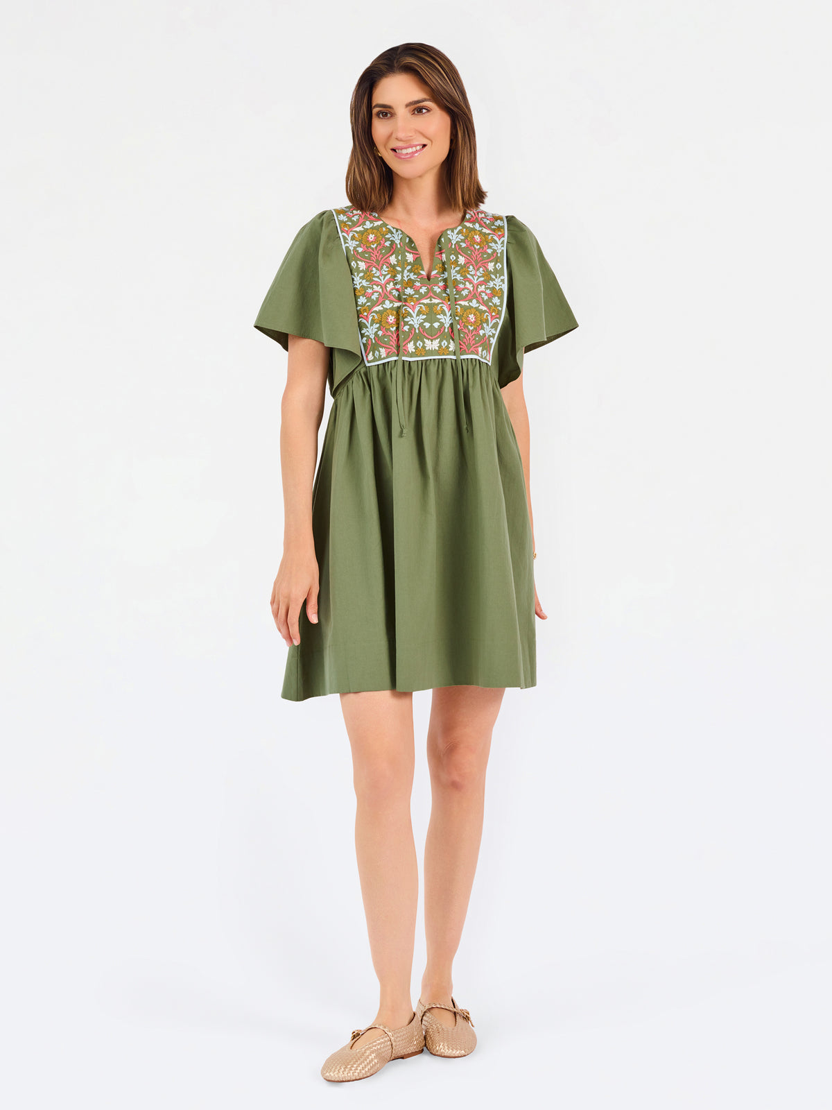 A woman modeling a short-sleeve olive green dress with a colorful floral embroidered yoke and a gathered waist, paired with beige flat shoes against a plain white background.