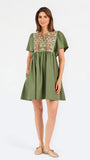 A woman modeling a short-sleeve olive green dress with a colorful floral embroidered yoke and a gathered waist, paired with beige flat shoes against a plain white background.