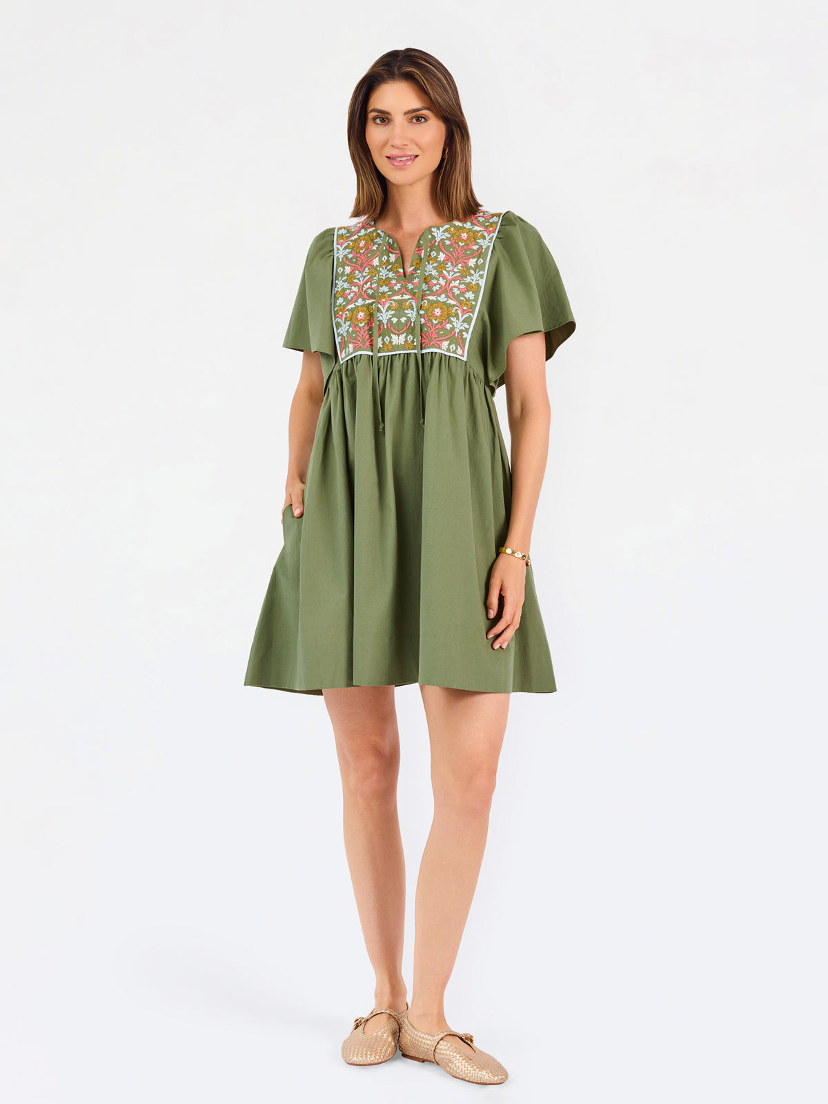 A woman modeling a short-sleeve olive green dress with a colorful floral embroidered yoke and a gathered waist, paired with beige flat shoes against a plain white background.