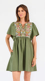 A woman modeling a short-sleeve olive green dress with a colorful floral embroidered yoke and a gathered waist, paired with beige flat shoes against a plain white background.