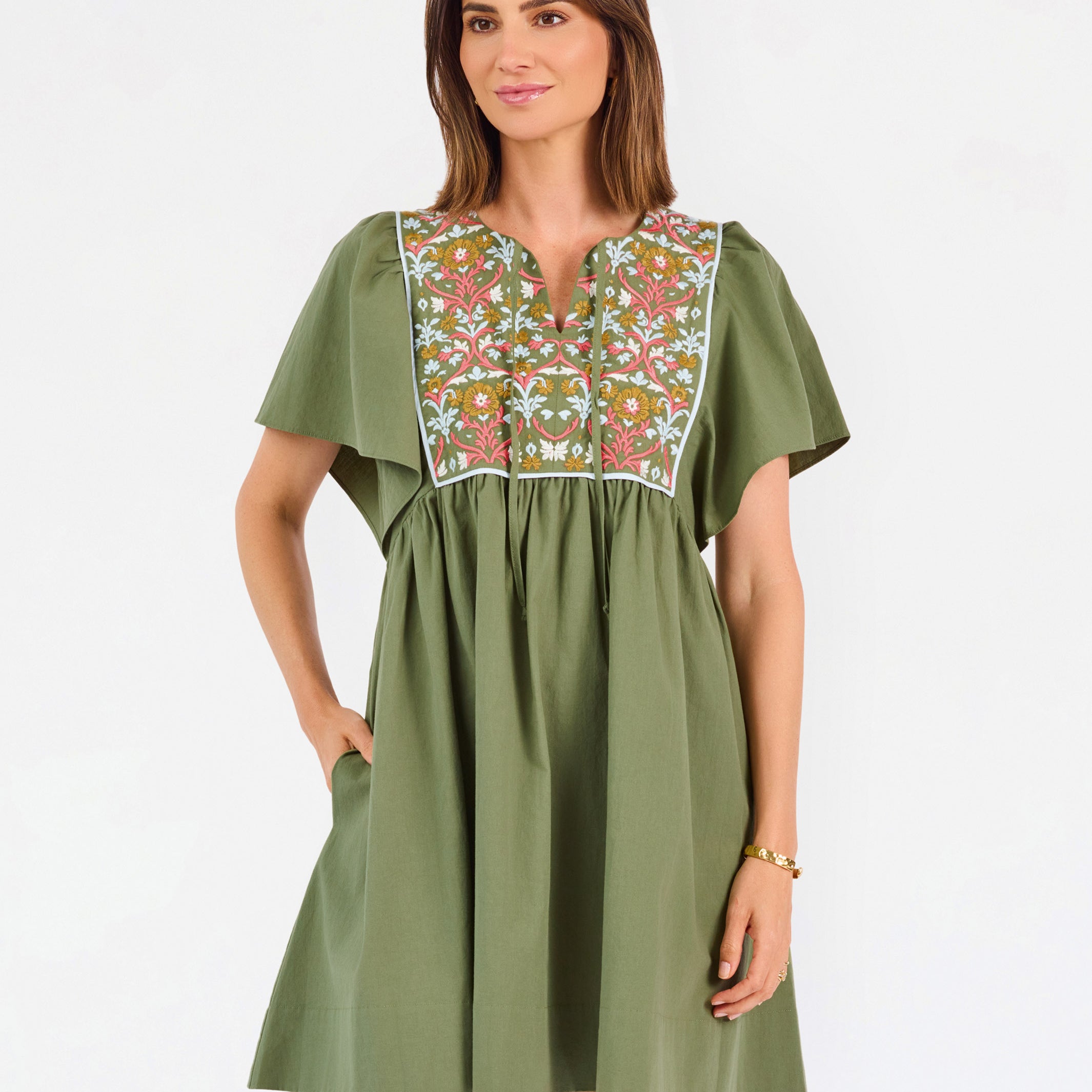 A woman modeling a short-sleeve olive green dress with a colorful floral embroidered yoke and a gathered waist, paired with beige flat shoes against a plain white background.