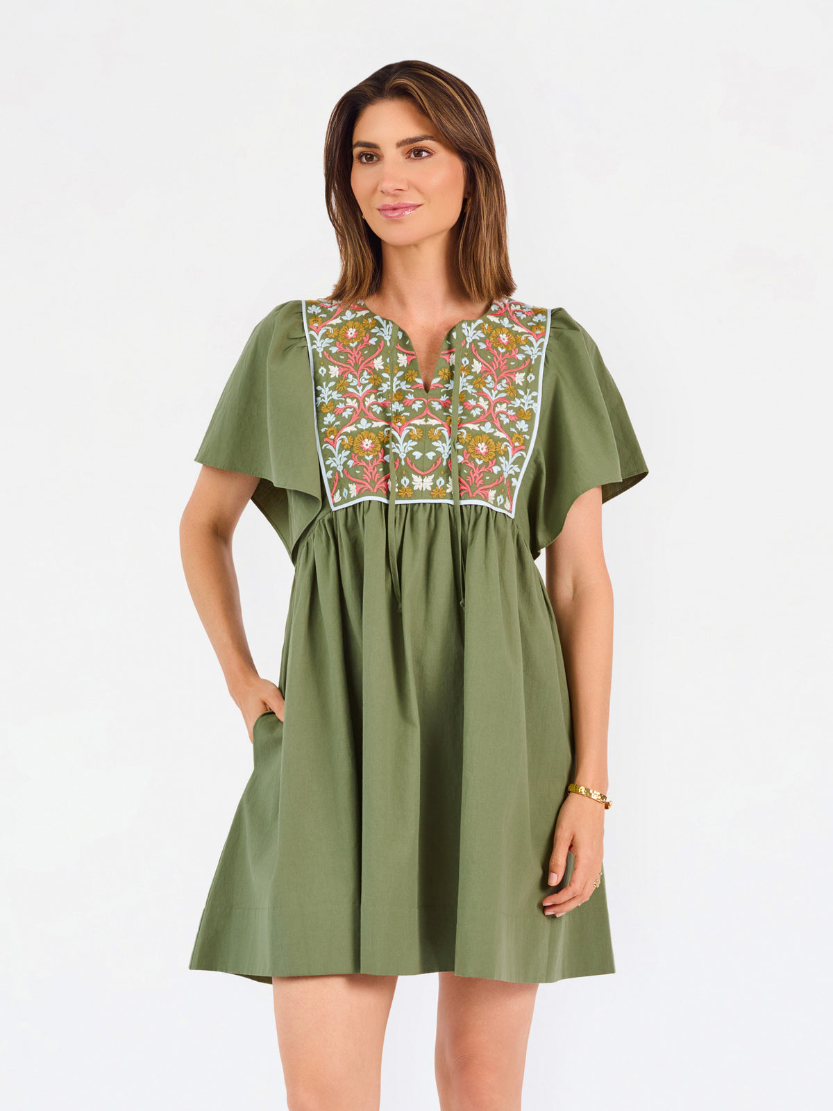 A woman modeling a short-sleeve olive green dress with a colorful floral embroidered yoke and a gathered waist, paired with beige flat shoes against a plain white background.