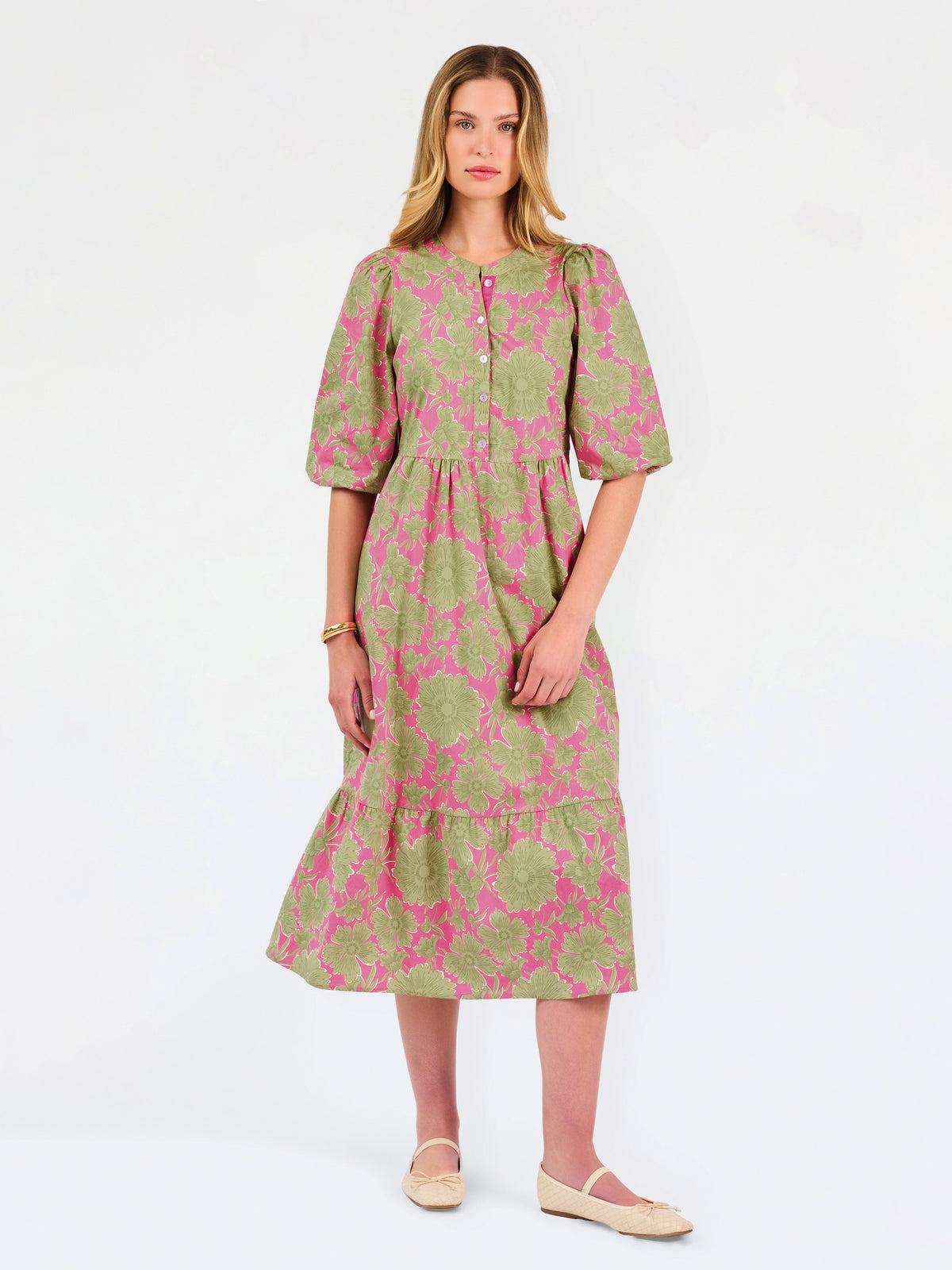 A woman modeling a midi dress with a green and pink floral pattern, short puff sleeves, and a gathered waist, set against a plain white studio background.