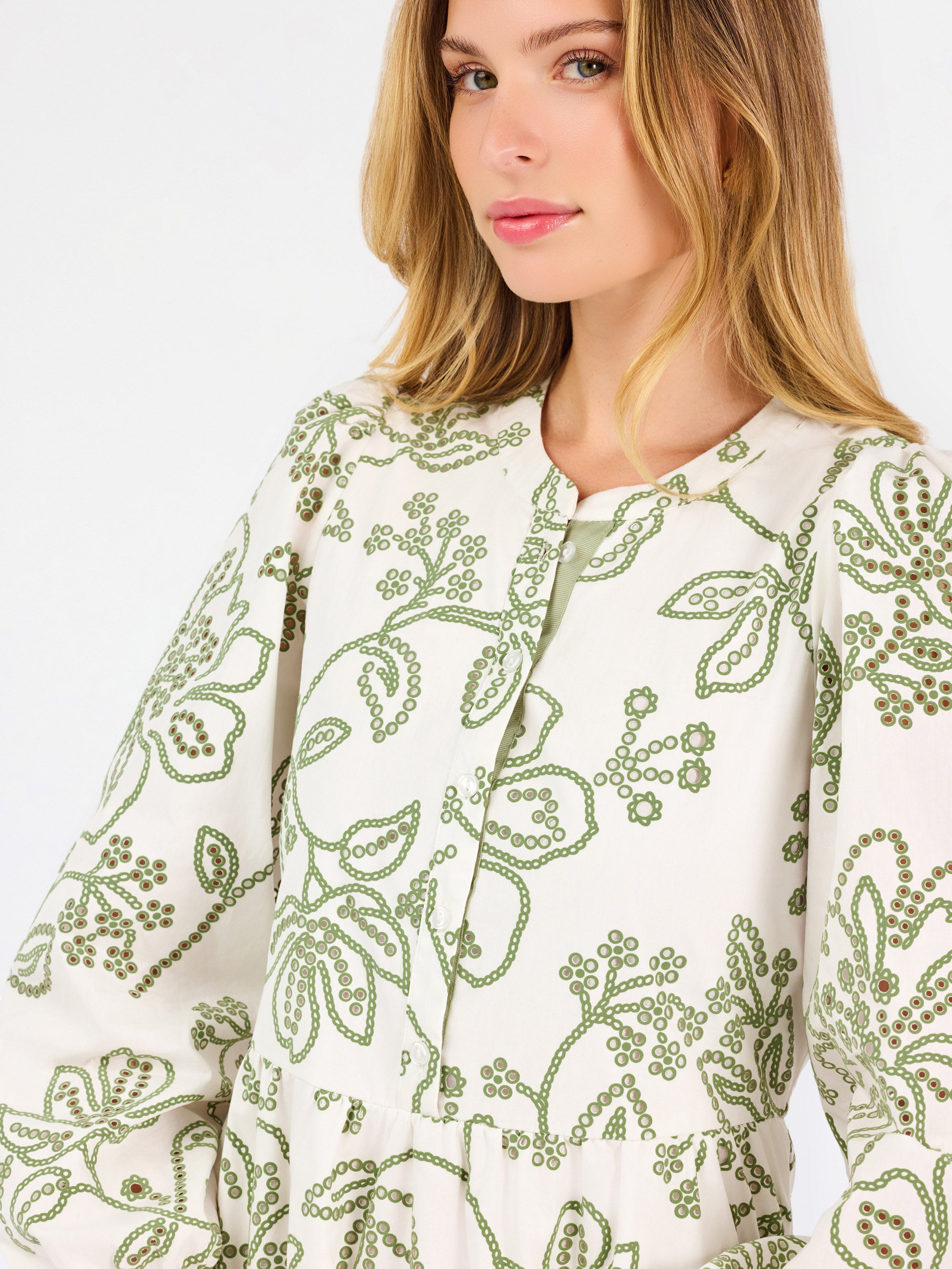 A close-up of a woman wearing a cream-colored long-sleeve dress with green floral embroidery and a keyhole neckline.