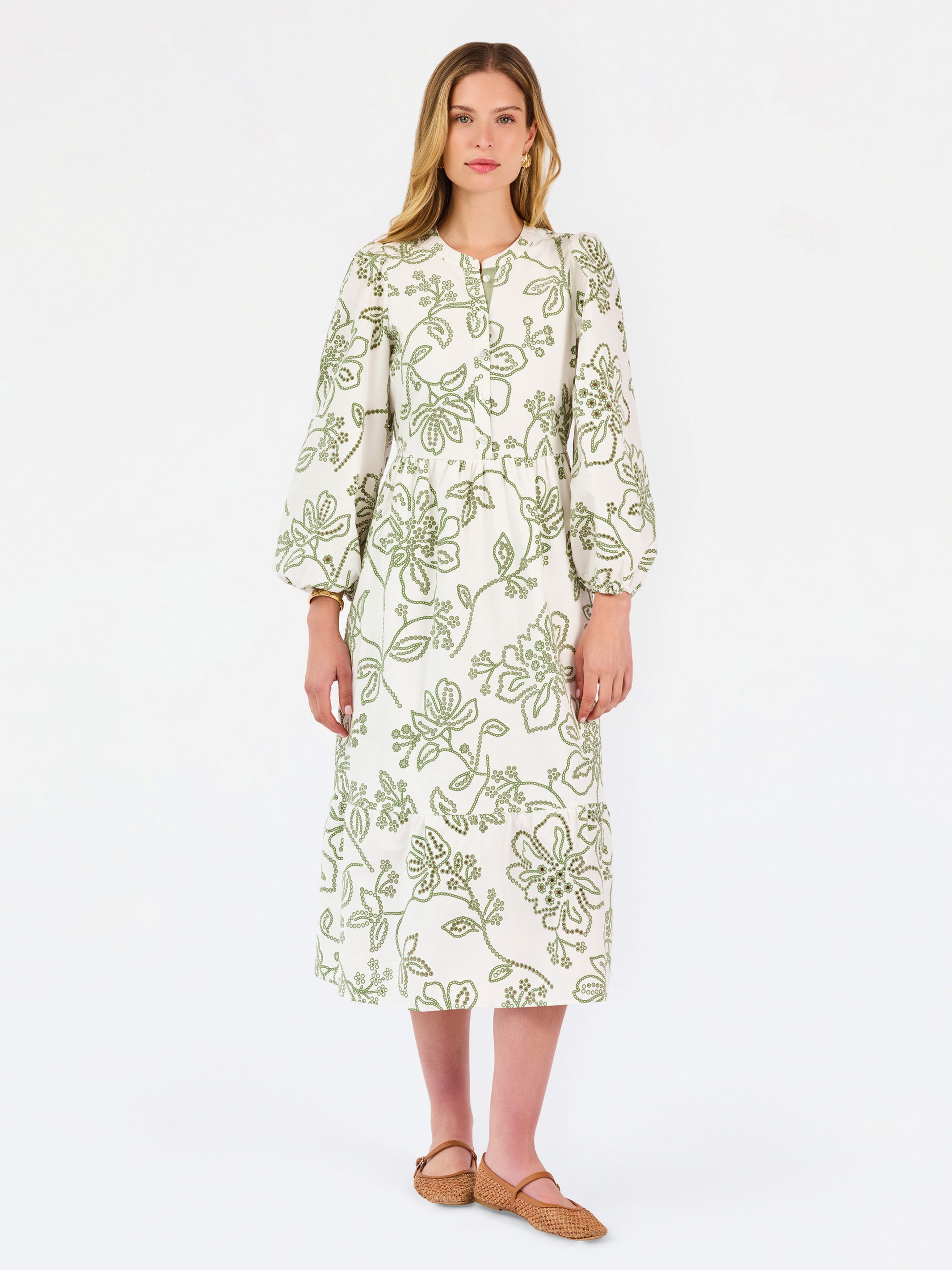 A woman modeling a long-sleeved midi dress with a green floral pattern on a cream background, paired with tan flat shoes, against a plain white studio background.