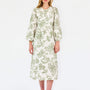 A woman modeling a long-sleeved midi dress with a green floral pattern on a cream background, paired with tan flat shoes, against a plain white studio background.