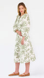 A woman modeling a long-sleeved midi dress with a green floral pattern on a cream background, paired with brown flat shoes, against a plain white studio background.