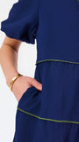 A woman modeling a navy blue tiered midi dress with short puff sleeves, a split neckline, and thin lime green contrast stitching, paired with beige flat shoes against a white studio background.