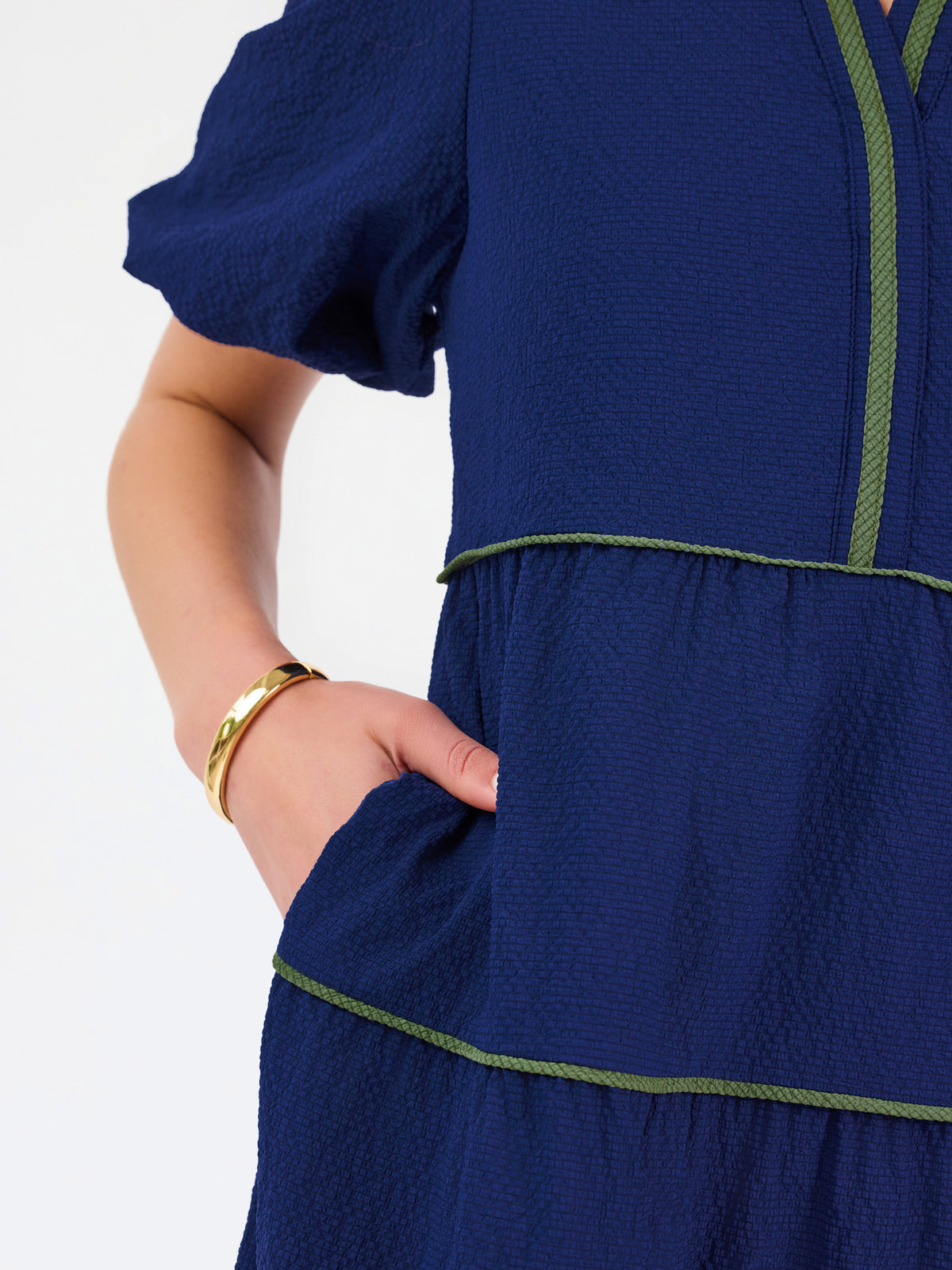 A woman modeling a navy blue tiered midi dress with short puff sleeves, a split neckline, and thin lime green contrast stitching, paired with beige flat shoes against a white studio background.