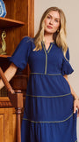 A woman wearing a royal blue tiered midi dress with gold trim, standing in a room with wooden bookshelves and a blue door frame.