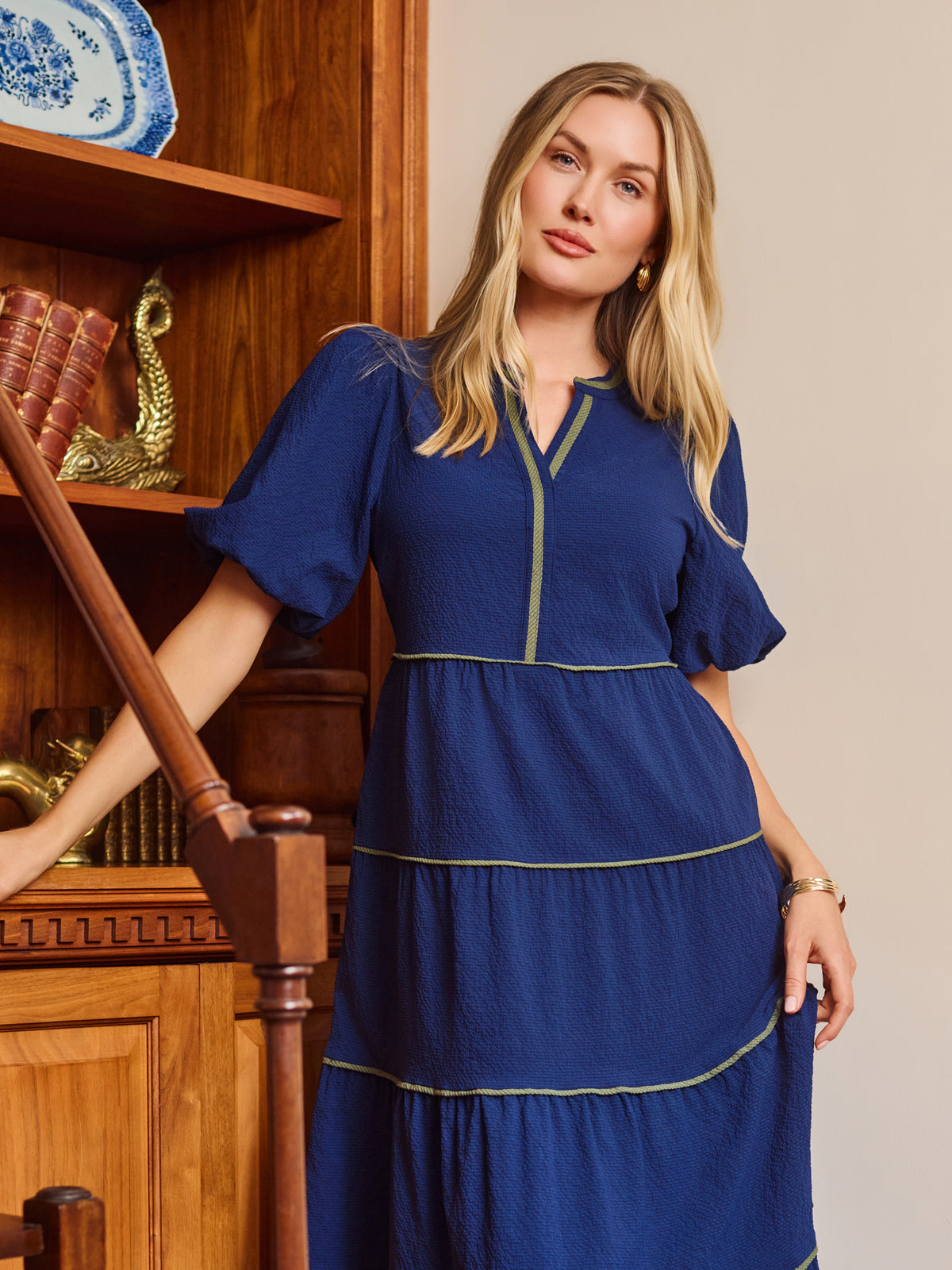 A woman wearing a royal blue tiered midi dress with gold trim, standing in a room with wooden bookshelves and a blue door frame.