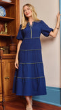 A woman wearing a royal blue tiered midi dress with gold trim, standing in a room with wooden bookshelves and a blue door frame.