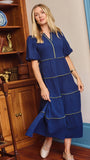 A woman wearing a royal blue tiered midi dress with gold trim, standing in a room with wooden bookshelves and a blue door frame.