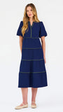 A woman modeling a navy blue tiered midi dress with short puff sleeves, a split neckline, and thin lime green contrast stitching, paired with beige flat shoes against a white studio background.