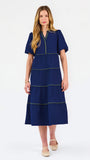 A woman modeling a navy blue tiered midi dress with short puff sleeves, a split neckline, and thin lime green contrast stitching, paired with beige flat shoes against a white studio background.