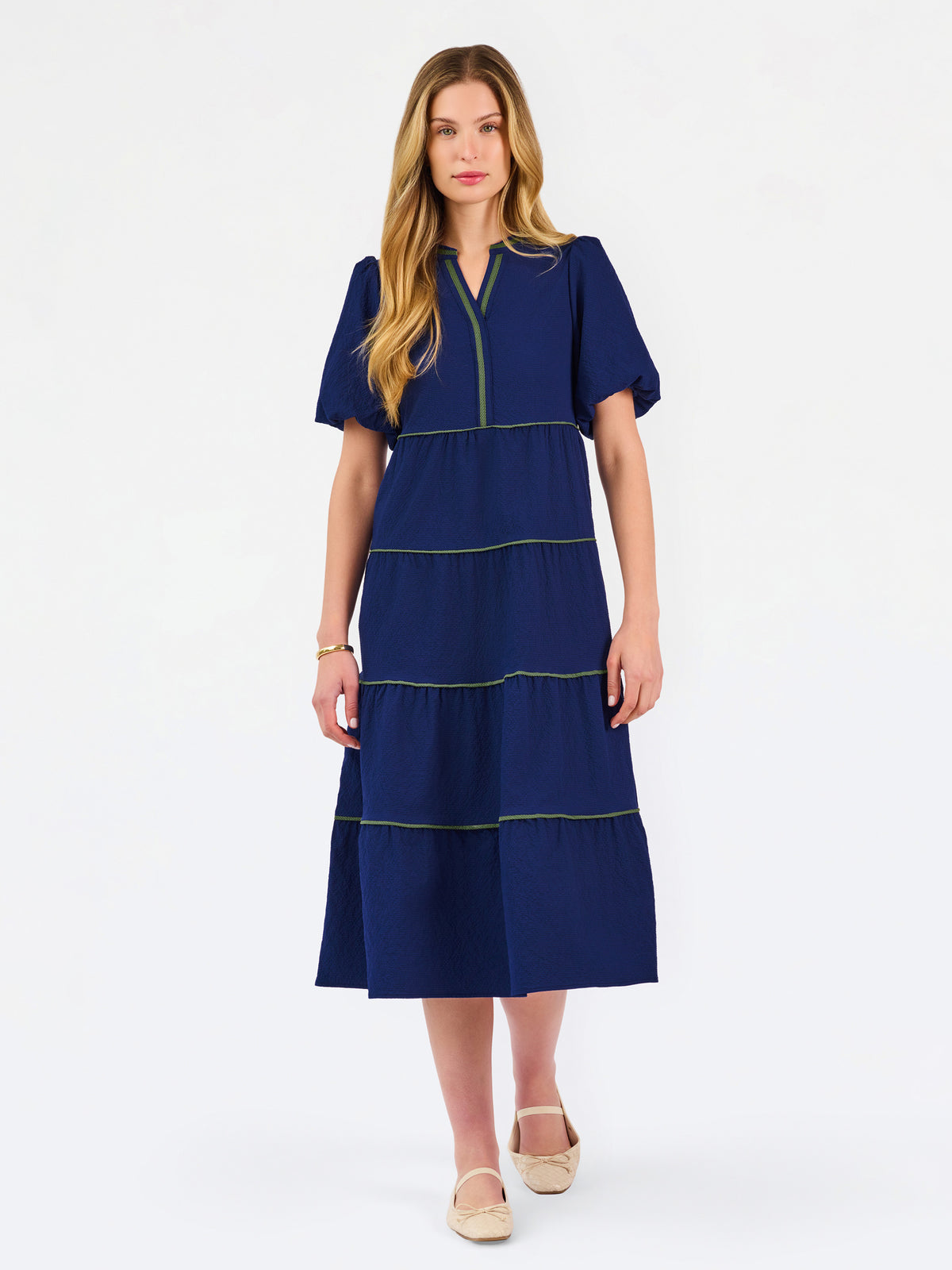 A woman modeling a navy blue tiered midi dress with short puff sleeves, a split neckline, and thin lime green contrast stitching, paired with beige flat shoes against a white studio background.