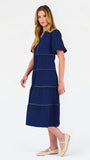 A woman modeling a navy blue tiered midi dress with short puff sleeves, a split neckline, and thin lime green contrast stitching, paired with beige flat shoes against a white studio background.