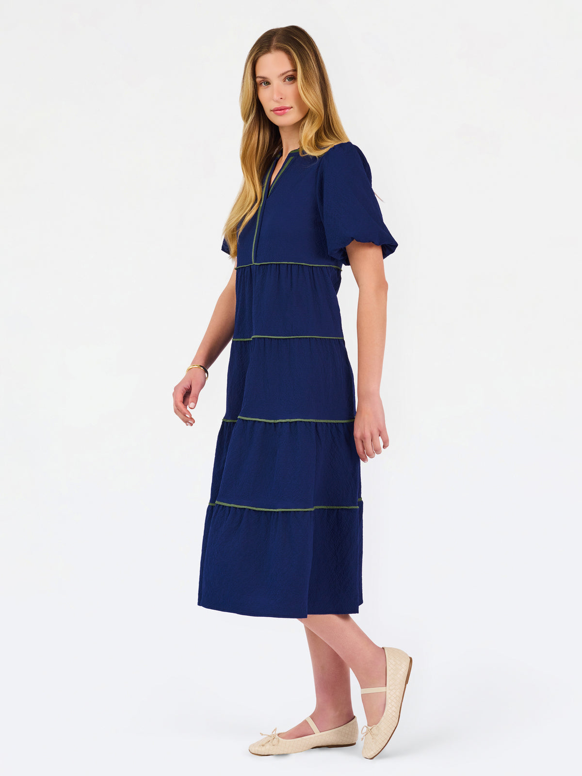 A woman modeling a navy blue tiered midi dress with short puff sleeves, a split neckline, and thin lime green contrast stitching, paired with beige flat shoes against a white studio background.