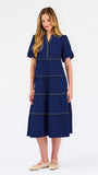 A woman modeling a navy blue tiered midi dress with short puff sleeves, a split neckline, and thin lime green contrast stitching, paired with beige flat shoes against a white studio background.