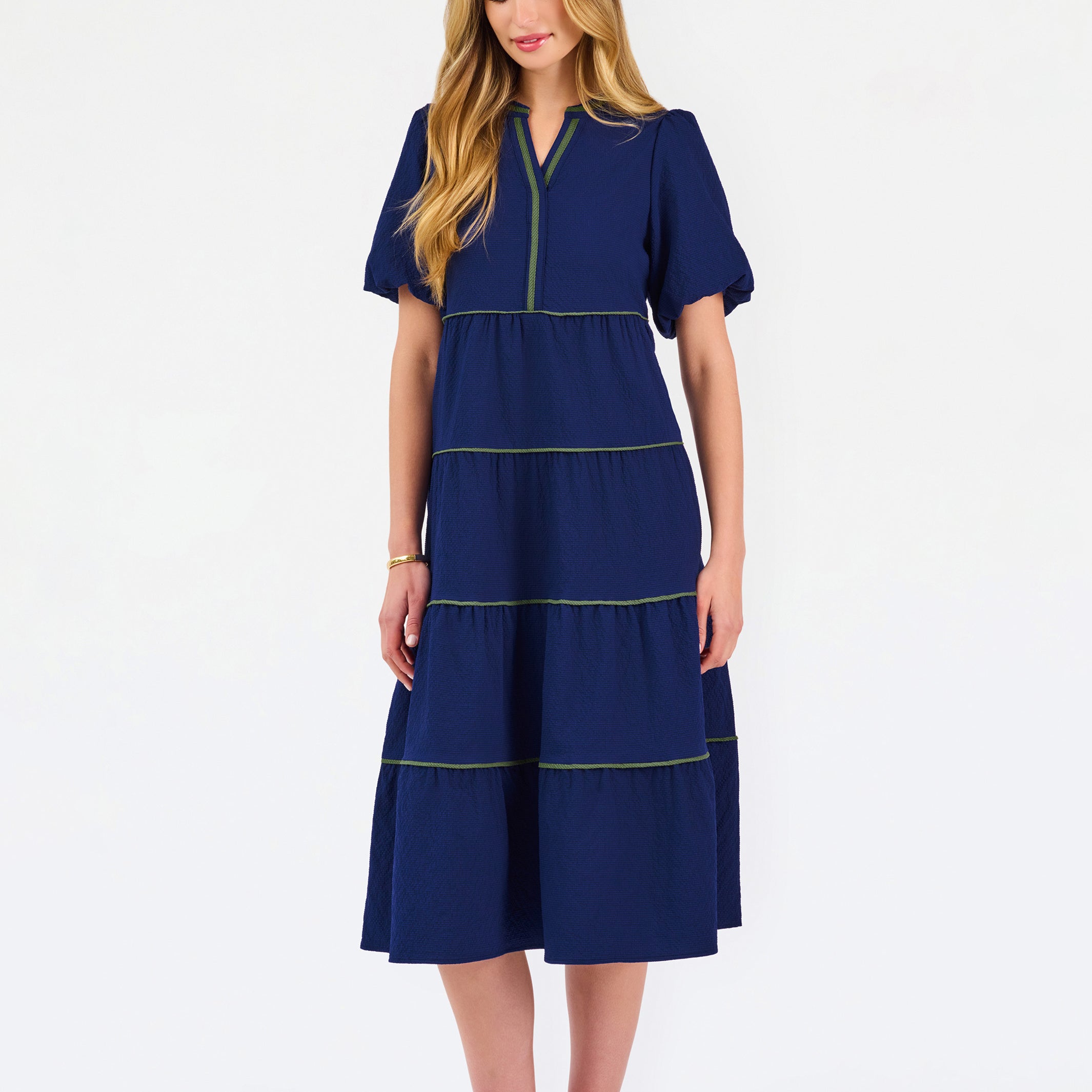 A woman modeling a navy blue tiered midi dress with short puff sleeves, a split neckline, and thin lime green contrast stitching, paired with beige flat shoes against a white studio background.
