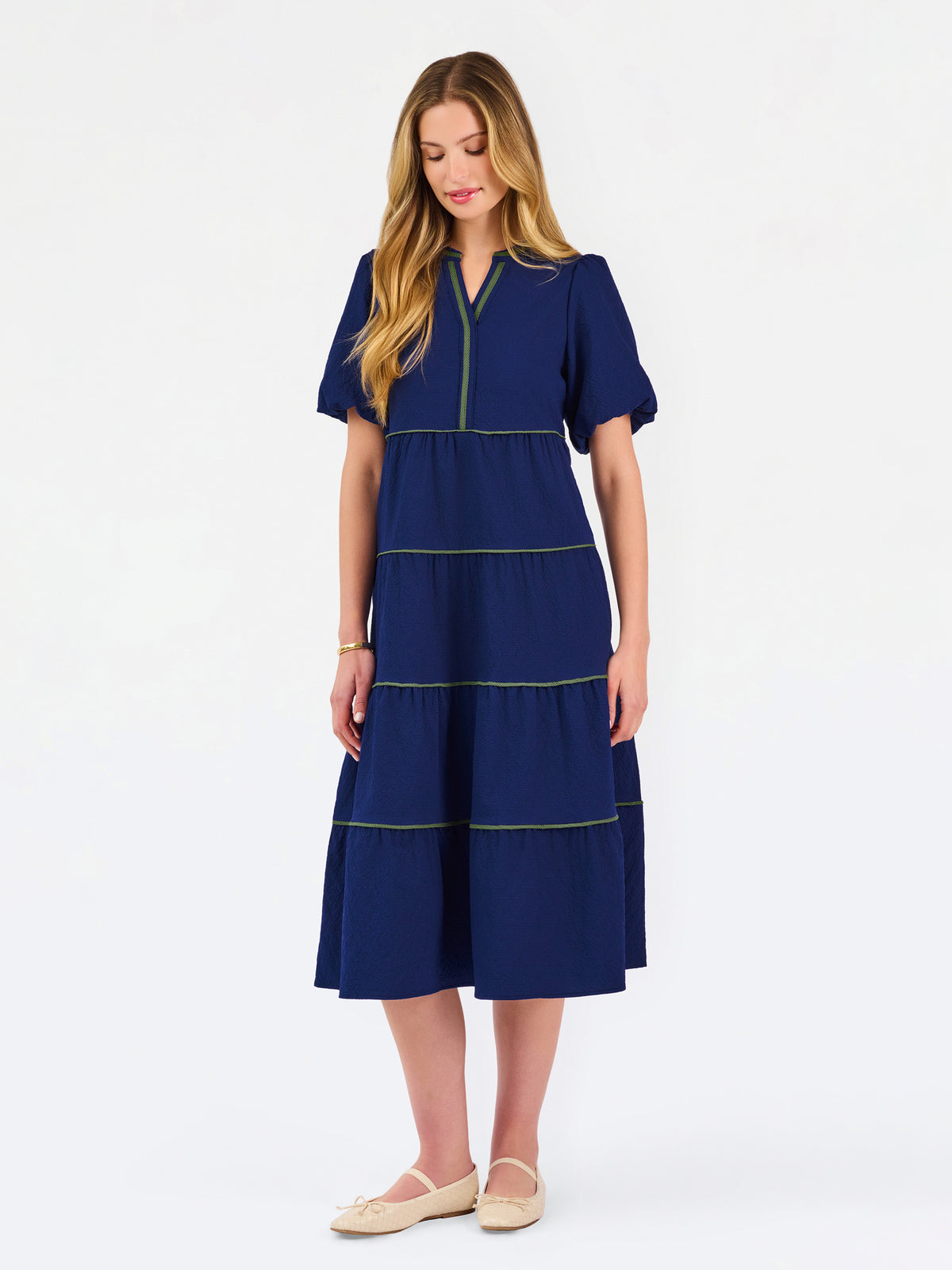 A woman modeling a navy blue tiered midi dress with short puff sleeves, a split neckline, and thin lime green contrast stitching, paired with beige flat shoes against a white studio background.