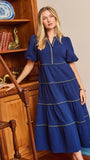 A woman wearing a royal blue tiered midi dress with gold trim, standing in a room with wooden bookshelves and a blue door frame.