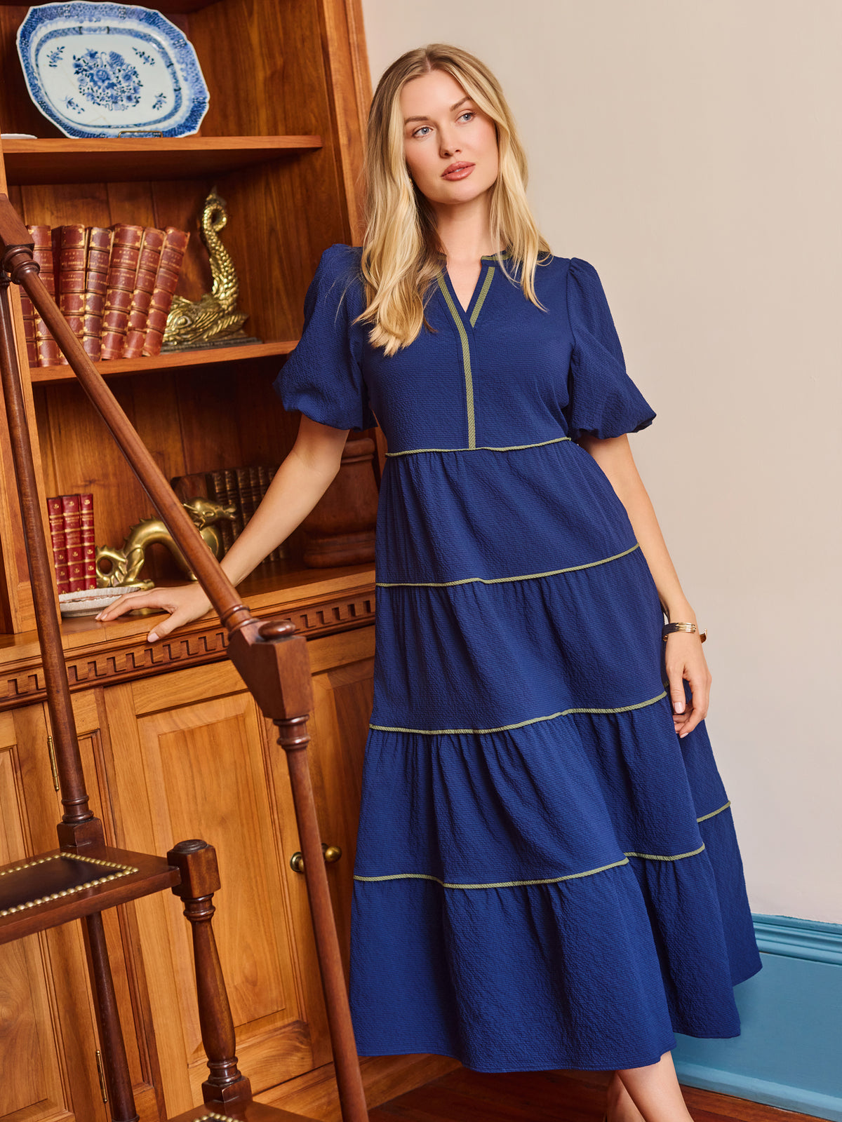 A woman wearing a royal blue tiered midi dress with gold trim, standing in a room with wooden bookshelves and a blue door frame.