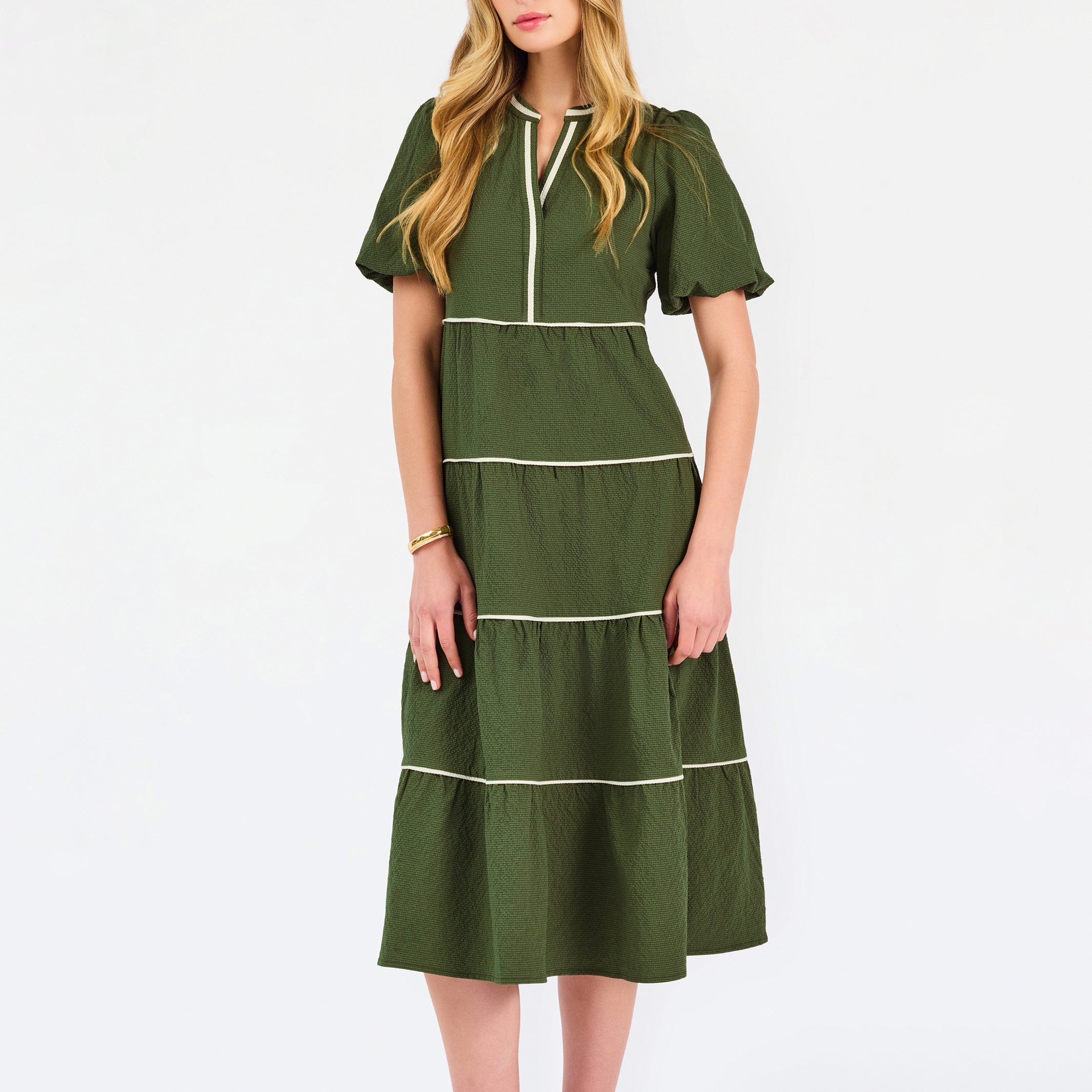 A woman modeling a short-sleeve olive green tiered midi dress with cream-colored piping details, paired with beige flat shoes, against a plain white studio background.