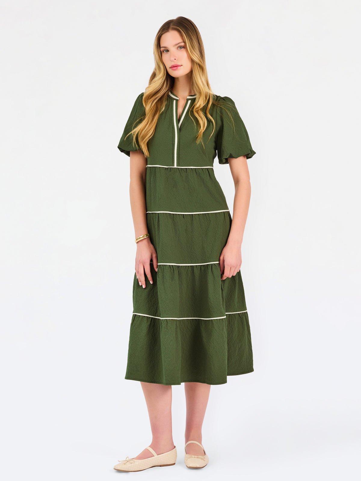A woman modeling a short-sleeve olive green tiered midi dress with cream-colored piping details, paired with beige flat shoes, against a plain white studio background.