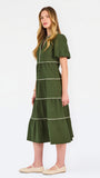 A woman modeling a short-sleeve olive green tiered midi dress with cream-colored piping details, paired with beige flat shoes, against a plain white studio background.