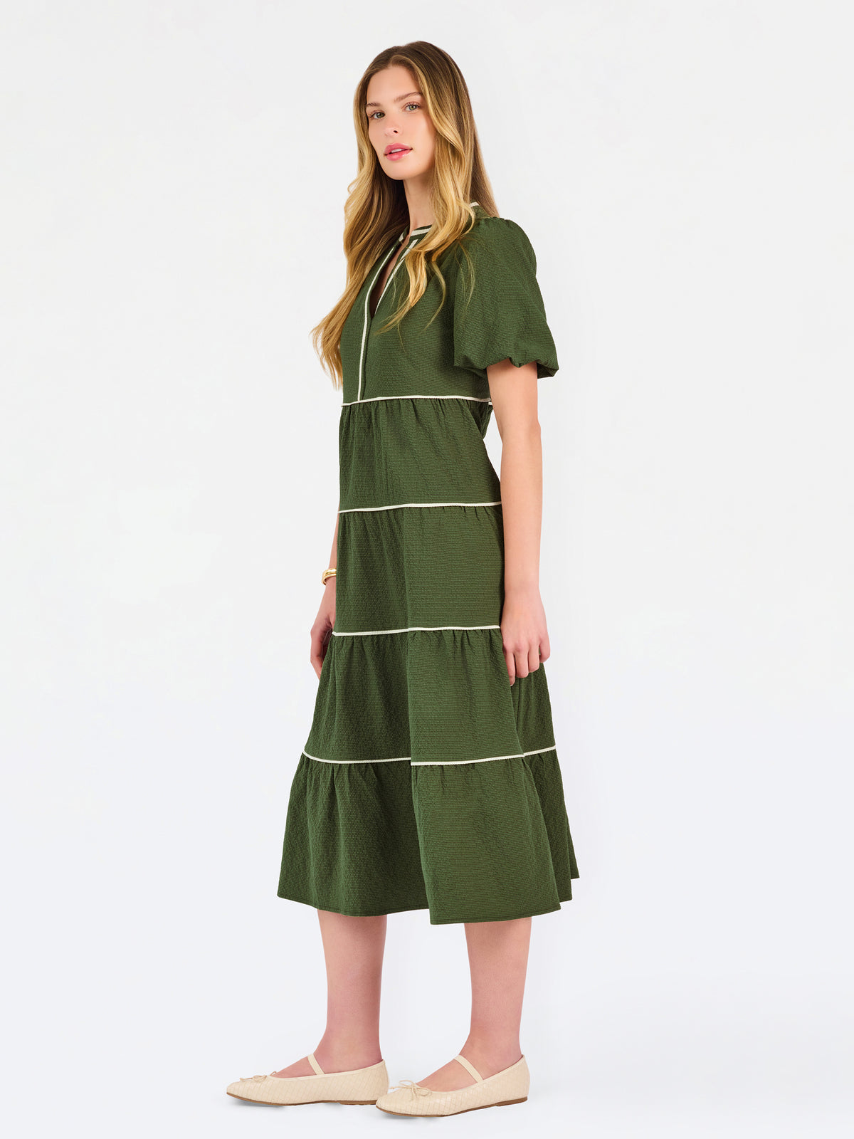 A woman modeling a short-sleeve olive green tiered midi dress with cream-colored piping details, paired with beige flat shoes, against a plain white studio background.