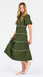 A woman modeling a short-sleeve olive green tiered midi dress with cream-colored piping details, paired with beige flat shoes, against a plain white studio background.