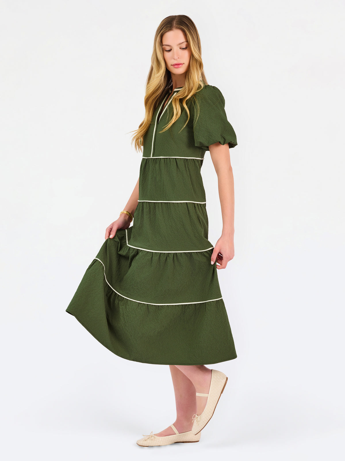A woman modeling a short-sleeve olive green tiered midi dress with cream-colored piping details, paired with beige flat shoes, against a plain white studio background.