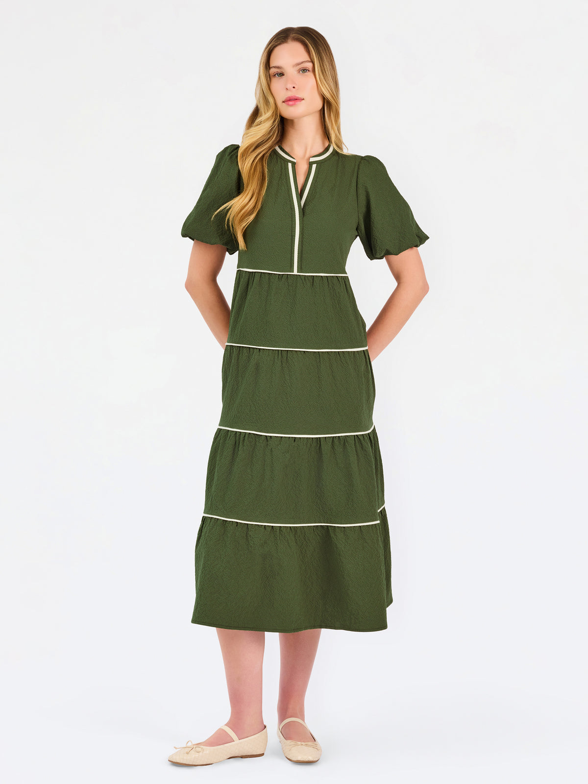 A woman modeling a short-sleeve olive green tiered midi dress with cream-colored piping details, paired with beige flat shoes, against a plain white studio background.