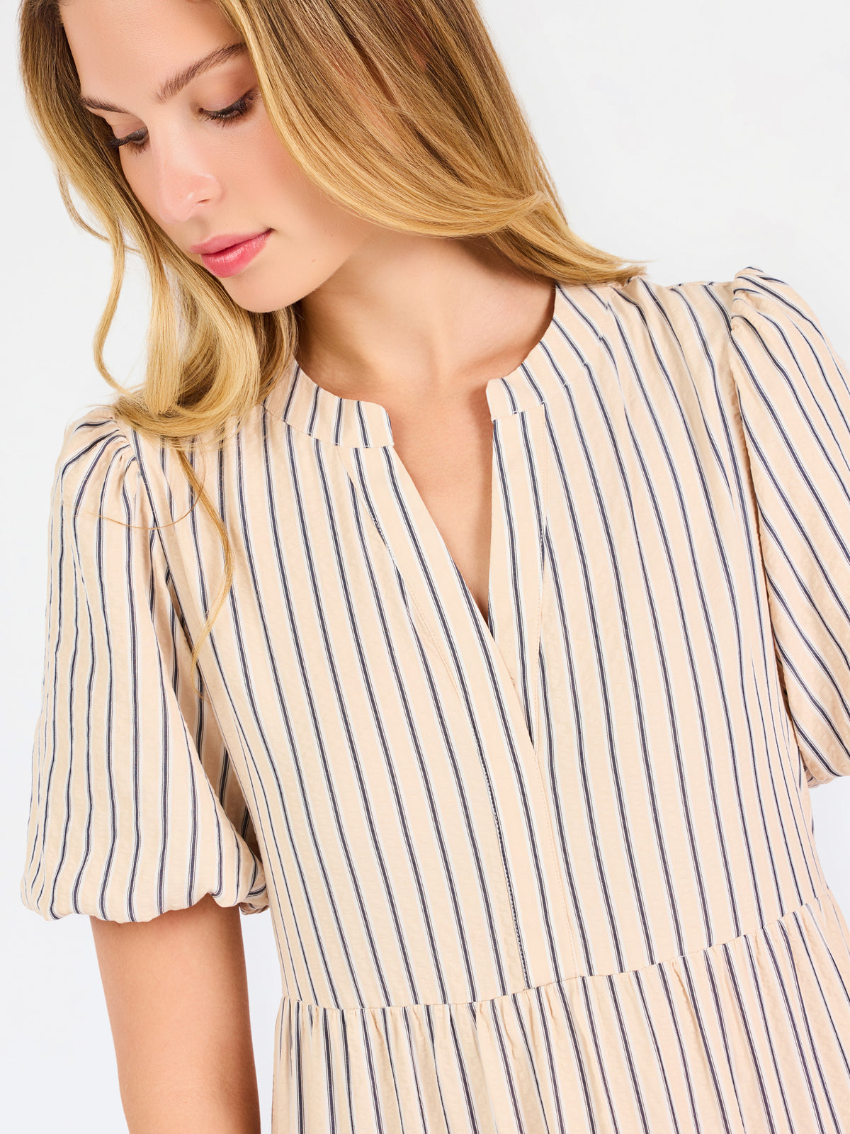 A woman modeling a short-sleeve midi dress with vertical beige and white stripes, featuring a tiered ruffle hem and a split neckline, set against a plain white studio background.