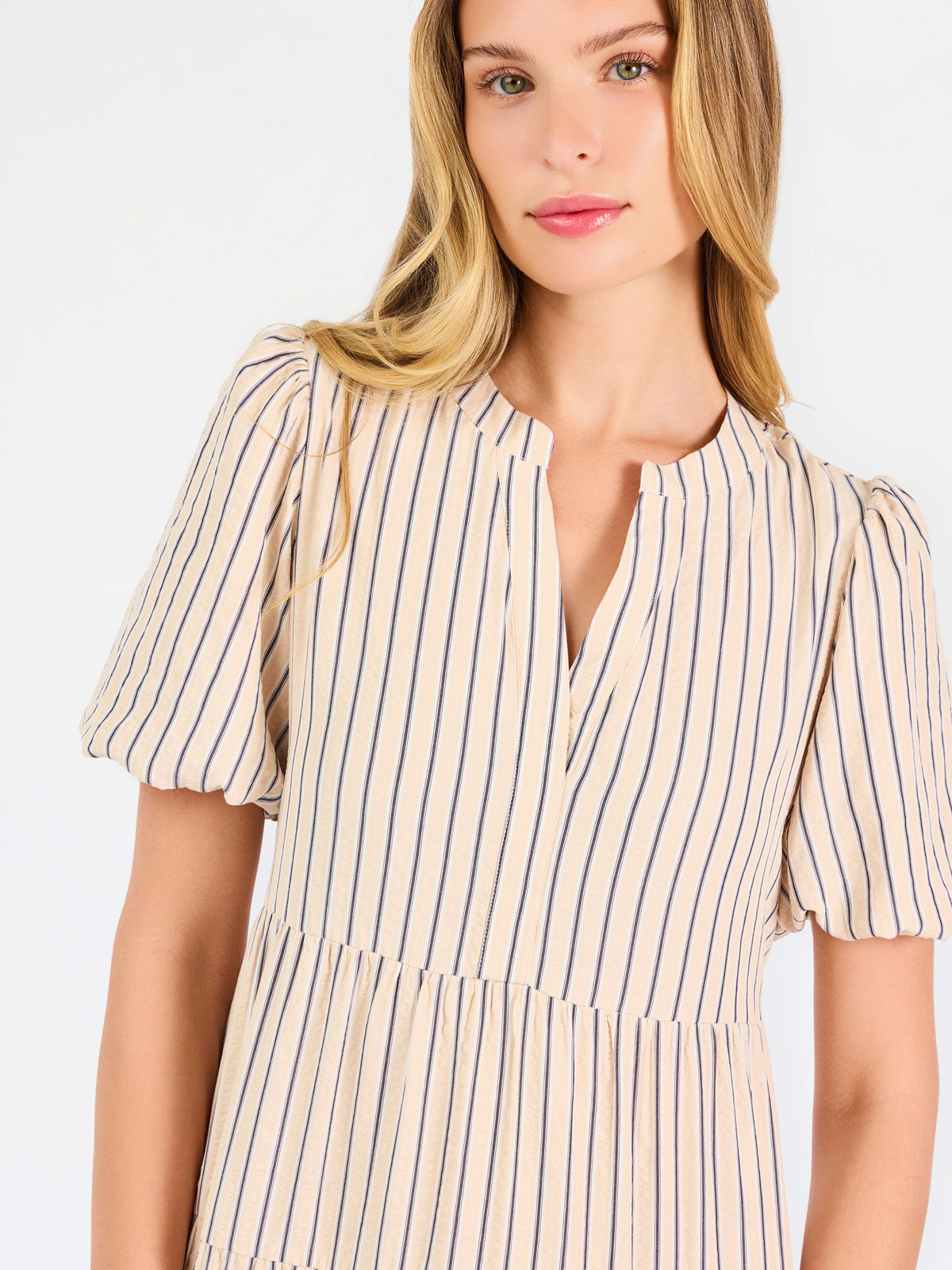 A woman modeling a short-sleeve midi dress with vertical beige and white stripes, featuring a tiered ruffle hem and a split neckline, set against a plain white studio background.