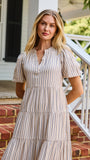 A woman stands on red brick steps in front of a white house porch, wearing a cream and tan vertically striped midi dress with puff sleeves and a tiered ruffle hem.