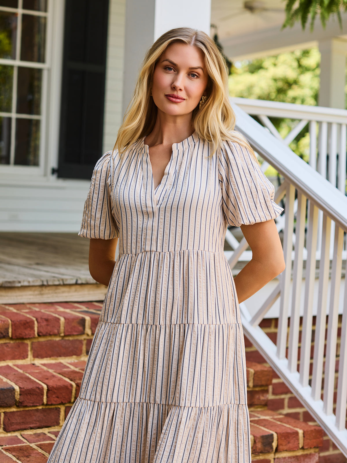 A woman stands on red brick steps in front of a white house porch, wearing a cream and tan vertically striped midi dress with puff sleeves and a tiered ruffle hem.